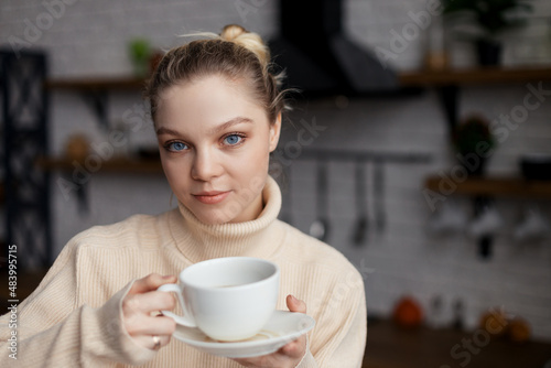 Happy young woman is in a cozy home atmosphere, sits at the kitchen table and holds a cup of coffee in her hands