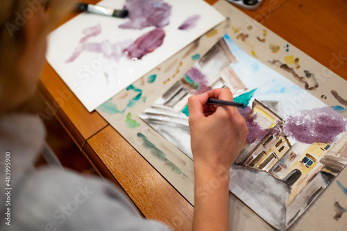 Brush in hand above watercolor painting. Colorful medieval city street on paper board.