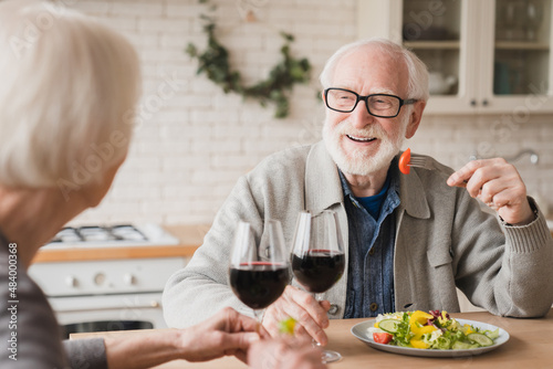 Love and relationship. Romantic date at home. Anniversary Valentine`s Day birthday celebration. Happy old elderly senior couple grandparents drinking wine having dinner at home kitchen together