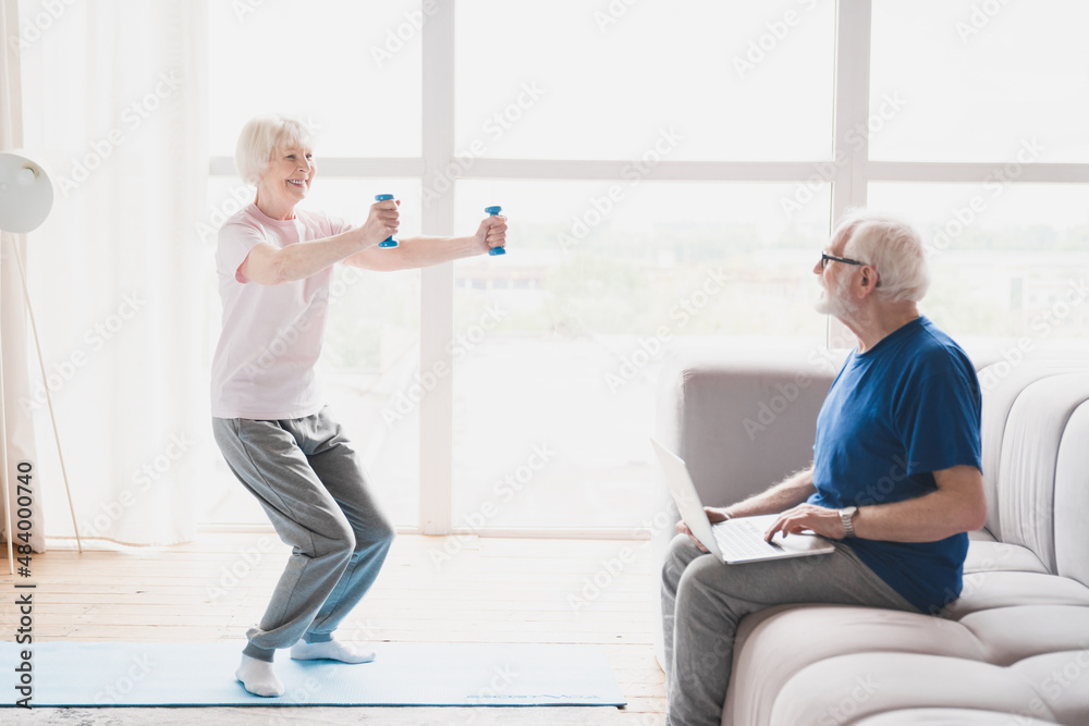 Fototapeta premium Old elderly senior grandparents training together, husband working using laptop while his wife stretching, doing physical exercises with dumbbells at home on lockdown