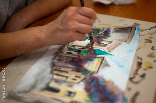 Brush in hand above watercolor painting. Colorful medieval city street on paper board.