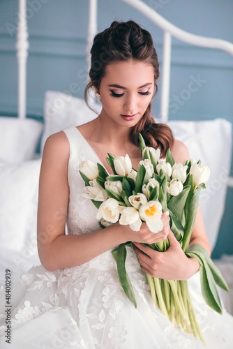 Beautiful bride with a wedding bouquet sitting on bed indoors in studio interior like at home. photo