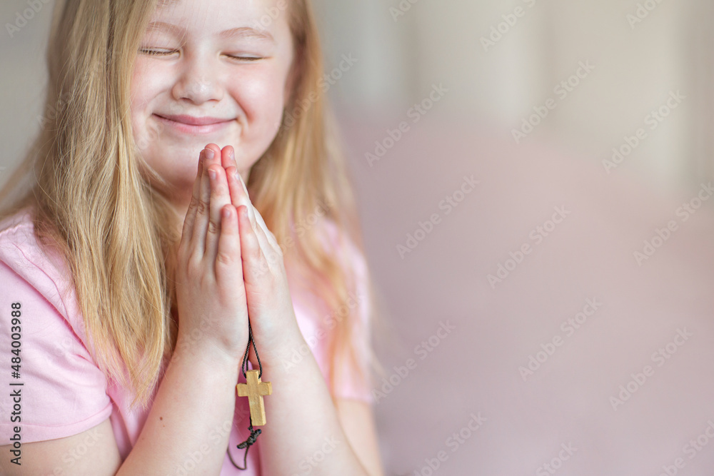 Naklejka premium A blonde little girl, smiling, praying at home in the morning with folded hands and holding a cross. Children's prayer, faith and prayer to God.