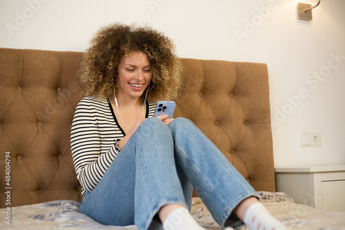 Happy young woman typing message on mobile phone and listening to favorite music in headphones. Cheerful white female with curly hair using modern smartphone gagdet for communication and entertainment photo