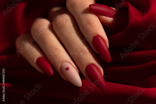 Fototapeta Matte red nails with small red heart on beige colour nail on the red fabric background