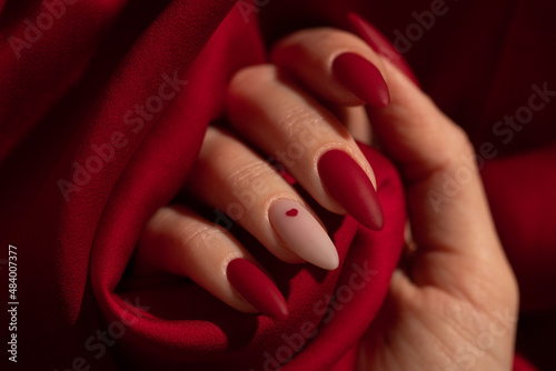 Fotografija Matte red nails with small red heart on beige colour nail on the red fabric background