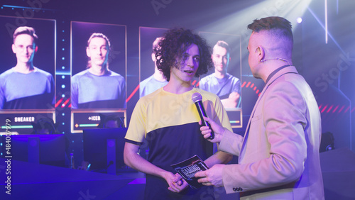 Young cyber sportsman smiling and speaking to adult male announcer while standing under neon illumination during gaming tournament photo