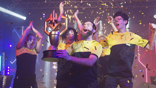 Bearded leader of cybersport team kissing golden trophy and dancing with team under confetti while celebrating victory in esports gaming championship