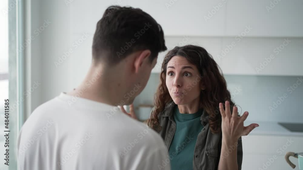 Married curly-haired woman quarrels angrily gesticulating and shouting with brunet husband over family problems standing in kitchen closeup Married curly-haired woman quarrels angrily gesticulating and shouting with brunet husband over family problems standing in kitchen closeup
