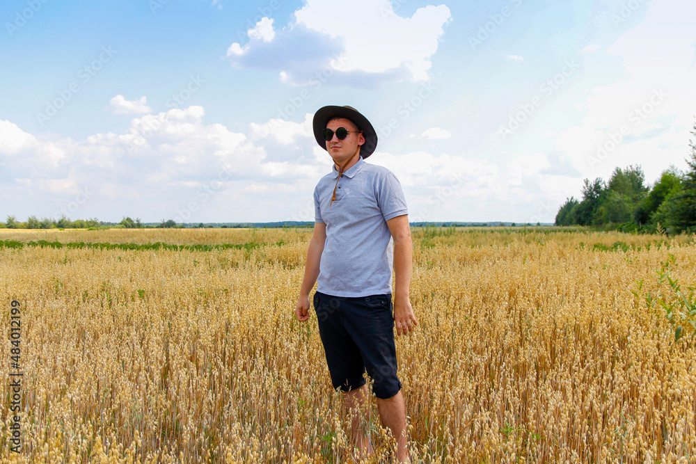 Crop and harvest. Portrait of farmer standing in gold wheat field with blue sky in background. Young man wearing sunglasses and cowboy hat in a field examining wheat crop. Oats plant