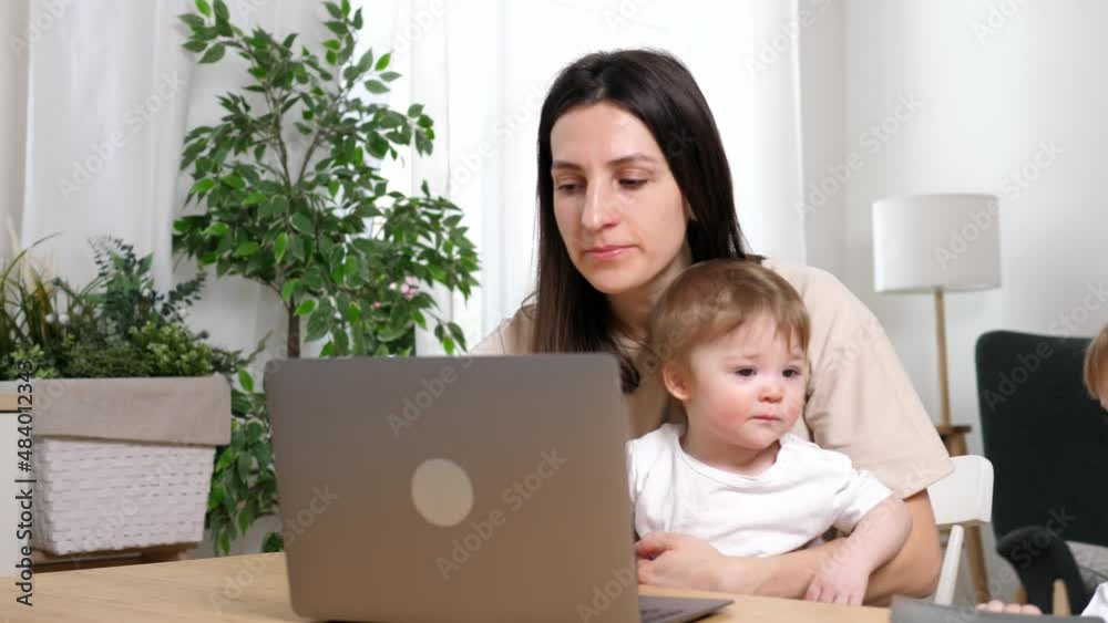 Happy and tired mother working from home, working at the table with a laptop, two sons are sitting in her arms. The concept of motherhood and parenting. Happy and tired mother working from home, working at the table with a laptop, two sons are sitting in her arms. The concept of motherhood and parenting.