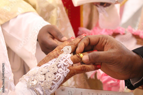 A closeup of an Indian groom hand putting the ring on a bride's finger in front of a prist photo
