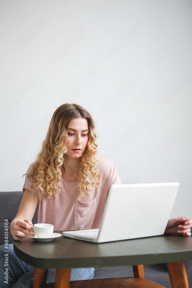 Beautiful young woman sitting on the sofa with white cup of coffee, working on white laptop. Girl in pink shirt looking to the monitor. Distance learning student Beautiful young woman sitting on the sofa with white cup of coffee, working on white laptop. Girl in pink shirt looking to the monitor. Distance learning student