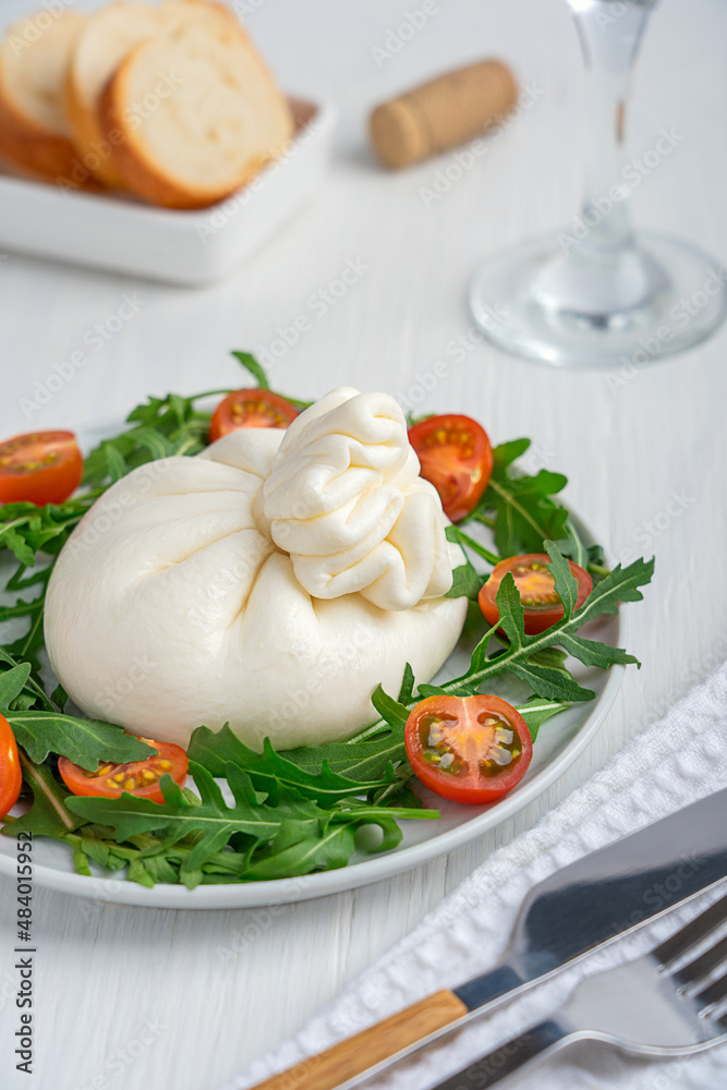 Homemade burrata Italian cow milk cheese with soft texture and buttery flavor served on plate with sliced cherry tomatoes and arugula leaves salad, bread and glass of wine on white wooden table Homemade burrata Italian cow milk cheese with soft texture and buttery flavor served on plate with sliced cherry tomatoes and arugula leaves salad, bread and glass of wine on white wooden table