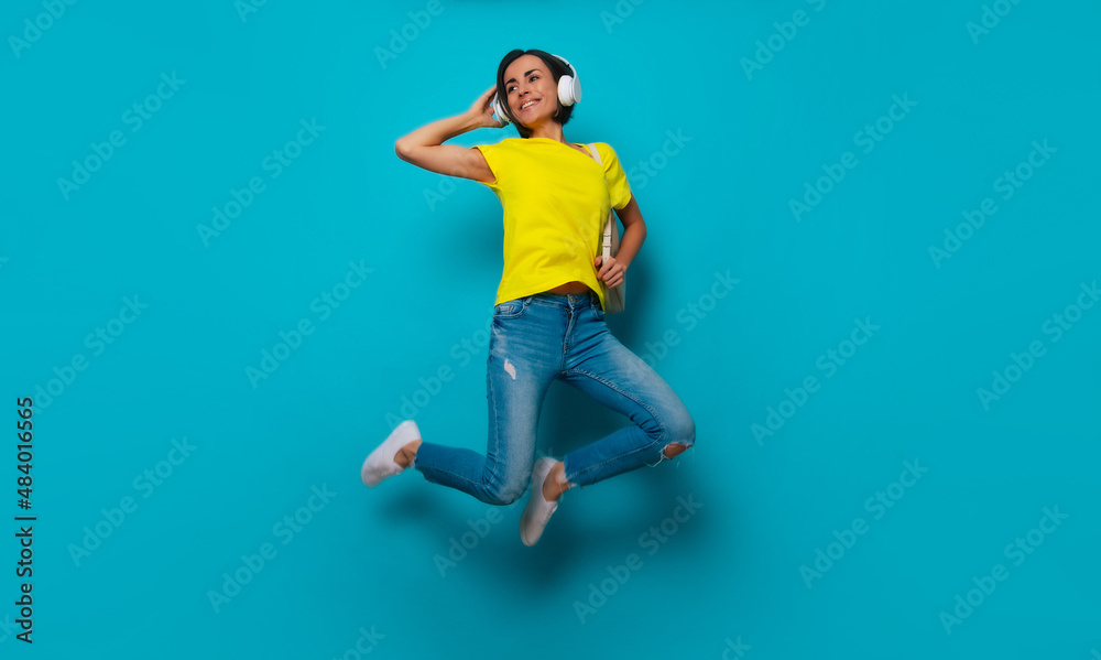 Full-length photo of excited attractive shouting modern woman in yellow t-shirt and headphones with backpack while she jumping isolated on blue background. Happy study process