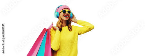Colorful portrait of stylish smiling young woman listening to music in headphones with shopping bags posing wearing a yellow knitted sweater, pink hat isolated on white background © rohappy