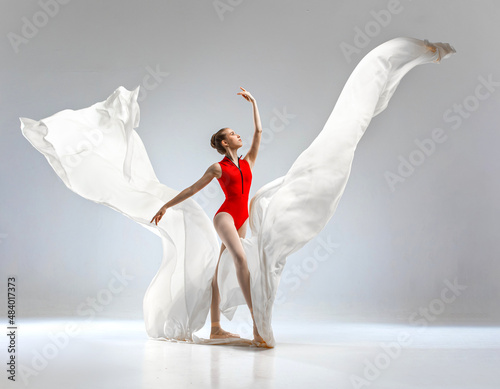 Fototapeta Naklejka Na Ścianę i Meble -  Ballerina in red ballet leotard dancing with white long cloth. She wears ballet pointe shoes. She dances in white studio