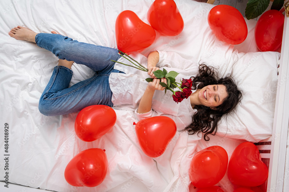 Excited Girl lying on bed red heart shape balloons with bouquet of flowers Woman rejoices gift on valentines day. Easy-going girl with long hair expressing positive emotions in valentines day.