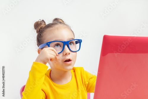 Portrait of beautiful cute little girl with glasses using laptop computer. Thoughtful nerd caucasian student studying online using notebook at home. Homeschooling, development and education concept. 