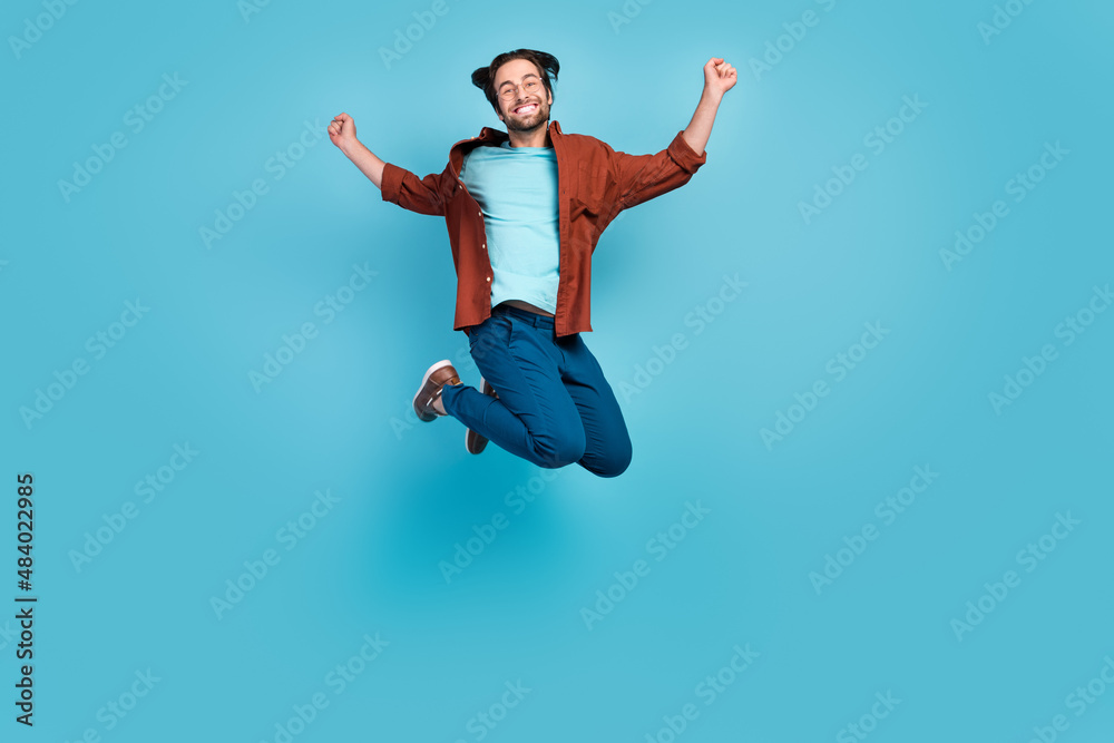 Full length photo of hooray brunet young guy jump wear eyewear shirt trousers sneakers isolated on blue background Full length photo of hooray brunet young guy jump wear eyewear shirt trousers sneakers isolated on blue background