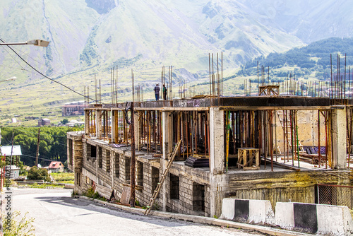 Multi-story concrete block building going up with lots of rebar and concrete blocks and two unrecognizable men standing on top and misty mountains of Kazbegi in background © Susan Vineyard 