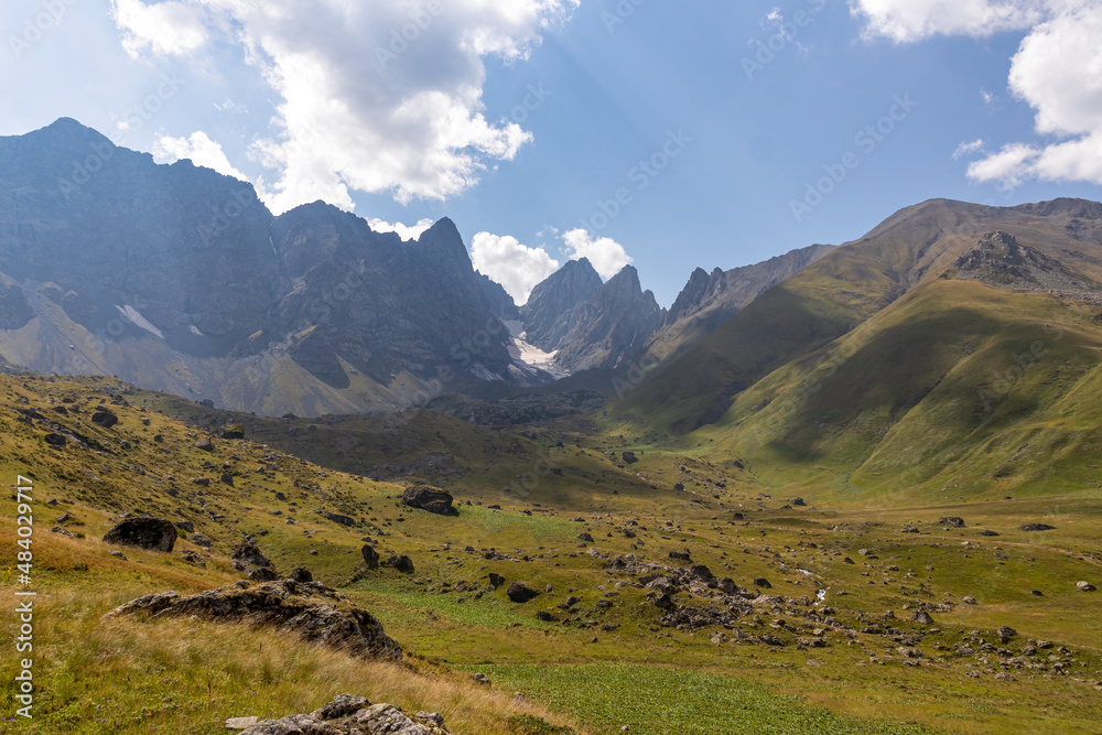 A panoramic view on the sharp mountain peaks of the Chaukhi massif in the Greater Caucasus Mountain Range in Georgia, Kazbegi Region. A hiking trail on a green alpine pasture. Georgian Dolomites.