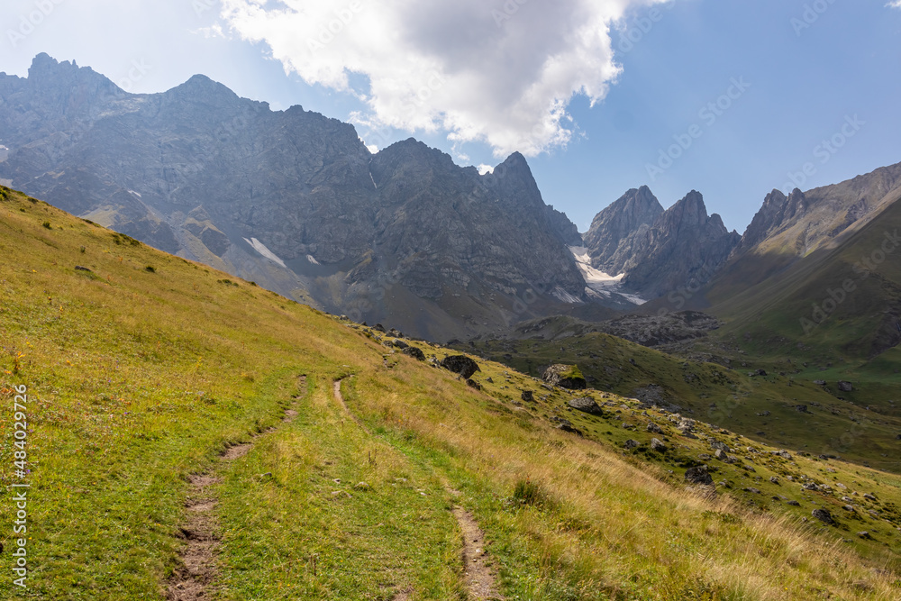 Fototapeta premium A panoramic view on the sharp mountain peaks of the Chaukhi massif in the Greater Caucasus Mountain Range in Georgia, Kazbegi Region. A hiking trail on a green alpine pasture. Georgian Dolomites.