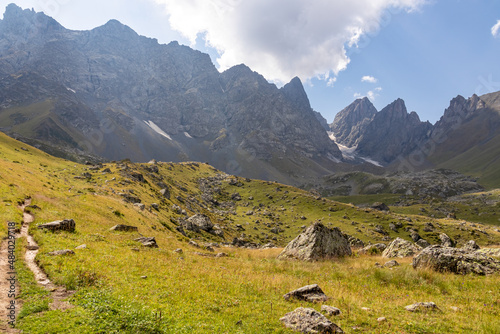 A panoramic view on the sharp mountain peaks of the Chaukhi massif in the Greater Caucasus Mountain Range in Georgia, Kazbegi Region. A hiking trail on a green alpine pasture. Georgian Dolomites.