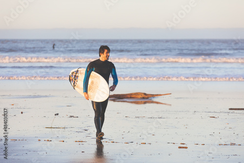Canvas Print Young caucasian man walking on the beach with a surfboard.