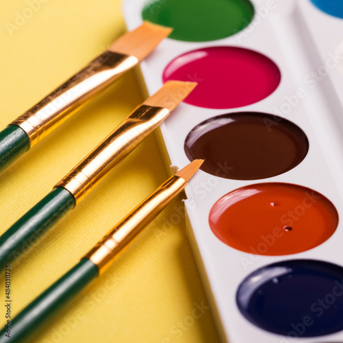 Multicolored watercolor paints with a brush on a yellow background. Objects for art