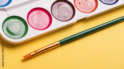 Multicolored watercolor paints with a brush on a yellow background. Objects for art