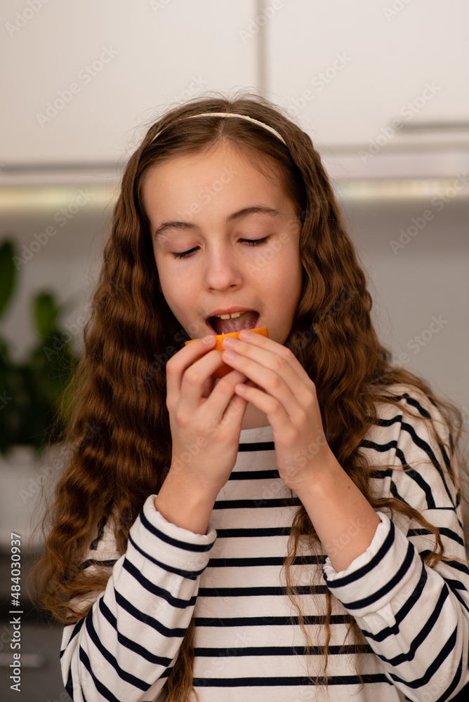 A sweet teenage girl eats an orange. She is dressed in a striped t-shirt. She's in the kitchen at home. Healthy eating. Family. Fresh fruit. A sweet teenage girl eats an orange. She is dressed in a striped t-shirt. She's in the kitchen at home. Healthy eating. Family. Fresh fruit.