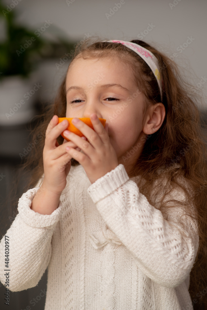 A sweet little girl eats an orange. She is dressed in white t-shirt. She's in the kitchen at home. Healthy eating. Family. Fresh fruit. A sweet little girl eats an orange. She is dressed in white t-shirt. She's in the kitchen at home. Healthy eating. Family. Fresh fruit.