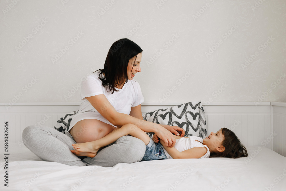 Pregnant mother plays with her little daughter in the bedroom on the bed. Mom and baby girl have fun together. Happy leisure time with family Pregnant mother plays with her little daughter in the bedroom on the bed. Mom and baby girl have fun together. Happy leisure time with family