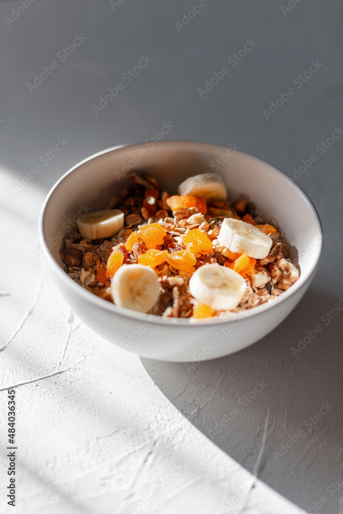Delicious healthy vegan raw breakfast with oatmeal, dried apricots, nuts and banana in a white bowl on the table in the early morning sun. Muesli food at sunrise