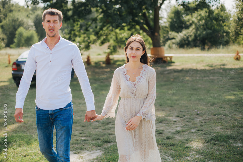 man and woman holding hands and walking on the green grass. Beautiful brunette in a long beige dress. Married couple on a walk man and woman holding hands and walking on the green grass. Beautiful brunette in a long beige dress. Married couple on a walk
