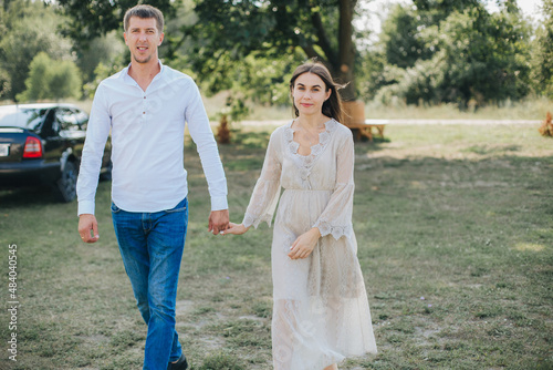 man and woman holding hands and walking on the green grass. Beautiful brunette in a long beige dress. Married couple on a walk
