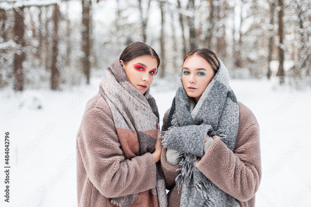 Two pretty young girls models with colored creative make-up in fashion winter clothes with vintage scarves and fur coats in a winter snowy forest. Russian female style