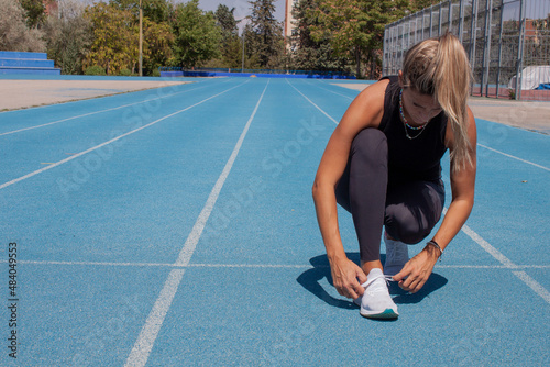 Woman on the running track tying the laces of her sneakers. Athletic training. Blue running track. Woman in black sportswear.