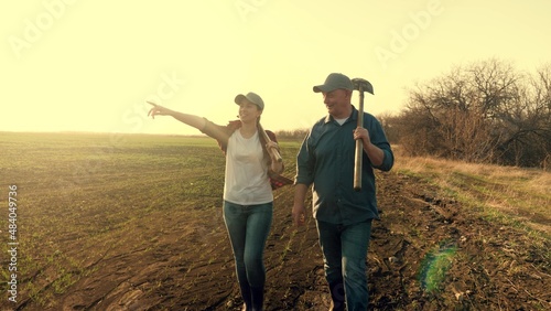 Farmers man and woman with shovel and chopper talk walking on cultivated field. People discuss hard working day issues passing bare trees at autumn sunset. Farmers man and woman talk walking
