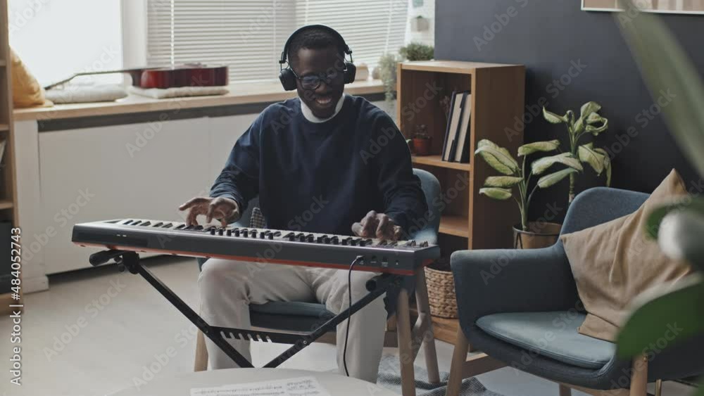 Medium long of happy Black man wearing eyeglasses and headphones, playing synthesizer in modern apartment at daytime, smiling