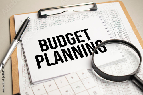 On the table are reports, a magnifying glass, a calculator, and a white notepad with the words BUDGET PLANNING. Business concept