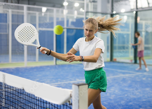 Teenage girl serving ball while playing padel in court during training.