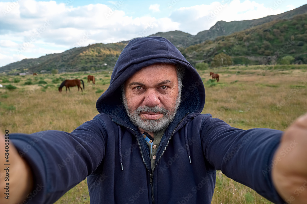 Happy, serious mature bearded man making a selfie on meadow with grazing horses. Concept of leisure activities,tourism, lifestyle e nature.