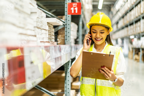 Asian woman in helmet and uniform talks on mobile phone discuss amount of stock product inventory on shelf at distribution warehouse factory. logistic business shipping and delivery service