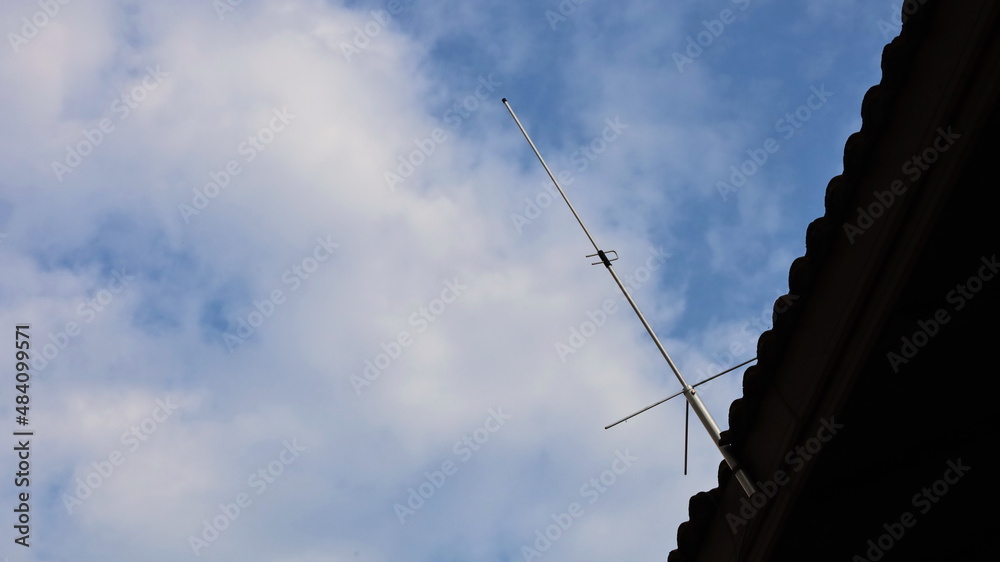 Communication antenna on the roof. Single Side Band (SSB) high frequency radio transmitting metal antenna on roof of house with blue sky background with white clouds with copy space. Selective focus Communication antenna on the roof. Single Side Band (SSB) high frequency radio transmitting metal antenna on roof of house with blue sky background with white clouds with copy space. Selective focus