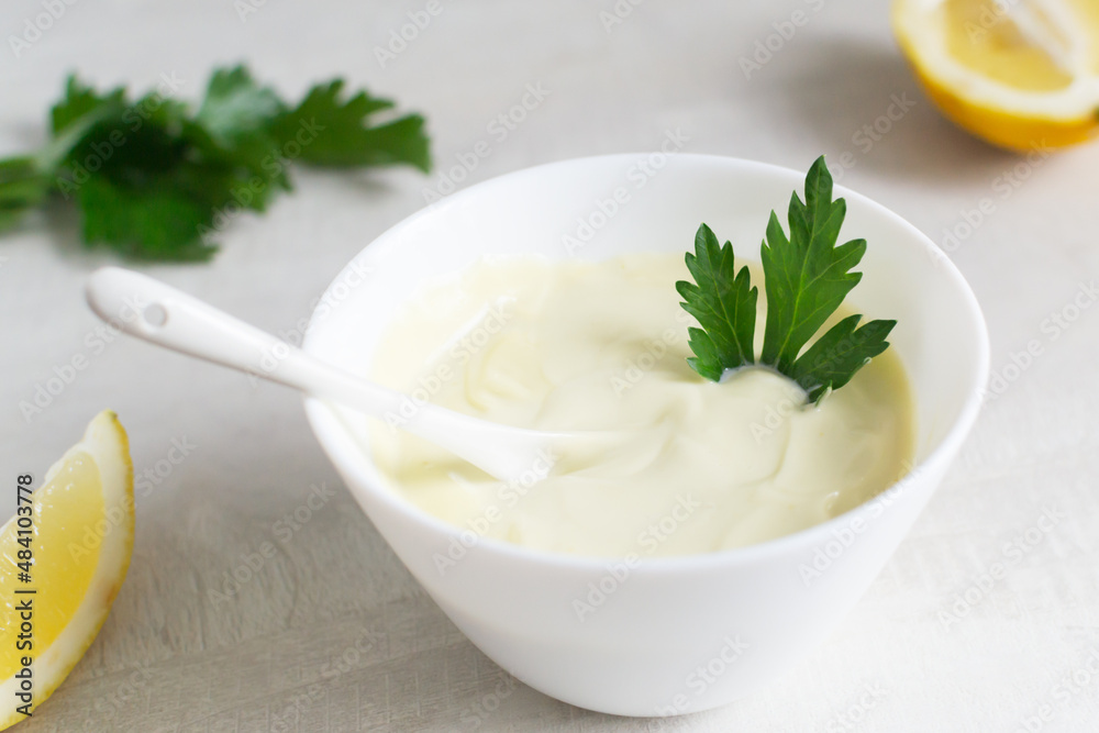 Homemade mayonnaise in a white bowl with parsley leaf and lemons in the background. The concept of food cooked at home. Selective focus. Vertical orientation. Homemade mayonnaise in a white bowl with parsley leaf and lemons in the background. The concept of food cooked at home. Selective focus. Vertical orientation.