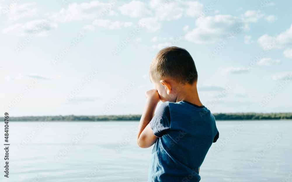 Little caucasian boy crying standing alone by water on shore outdoors on sunny summer day. Lonely sad, upset child in nature, back view Little caucasian boy crying standing alone by water on shore outdoors on sunny summer day. Lonely sad, upset child in nature, back view