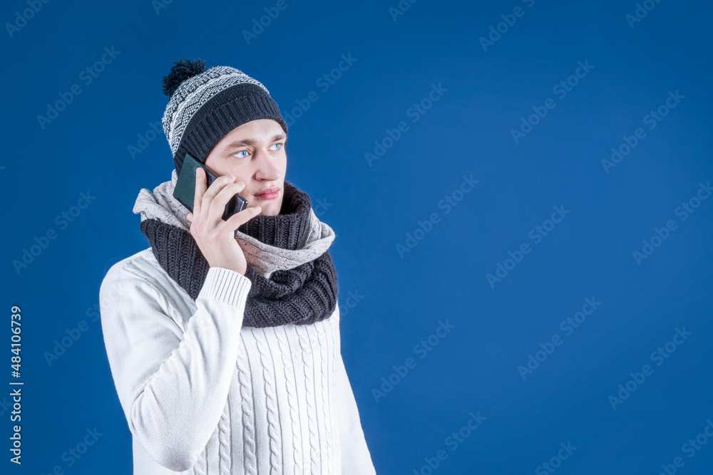 A man in a winter hat and a scarf is talking on the phone. The guy holds a smartphone in his hands. Conversation on a mobile phone. Winter. Coldly. A man in a winter hat and a scarf is talking on the phone. The guy holds a smartphone in his hands. Conversation on a mobile phone. Winter. Coldly.