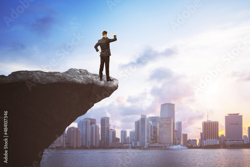 Attractive young european man on cliff edge looking into the distance on bright city background with daylight and mock up place. Future  success  leadership and career growth concept.